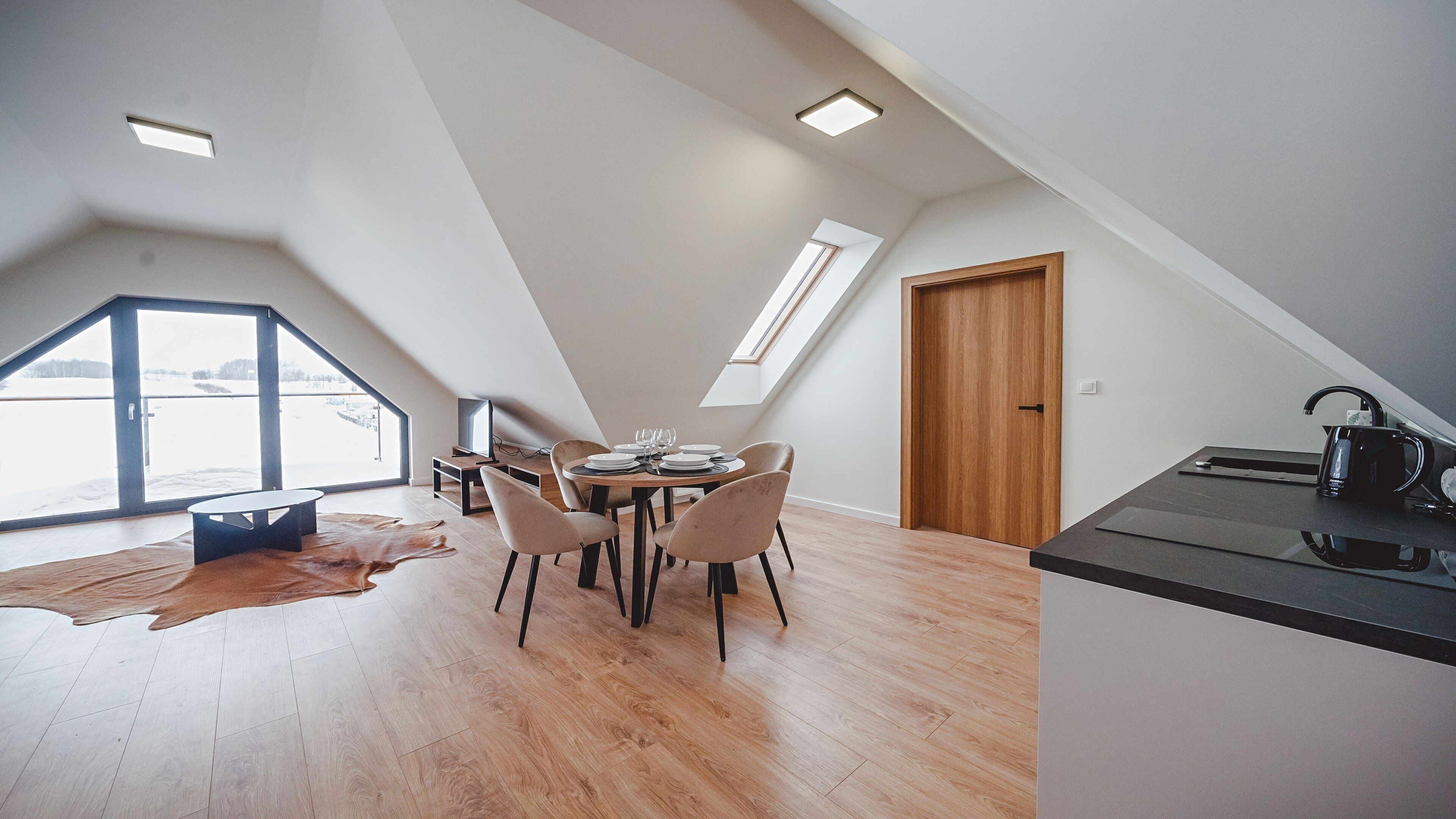 A modern loft with a dining area, a kitchen, and a large window overlooking a snowy landscape.
