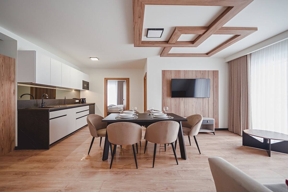 A modern hotel room with a dining area, a kitchen, and a TV mounted on the wall.