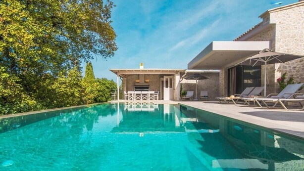 Ein klares Schwimmbecken mit Liegestühlen und einer überdachten Sitzgruppe, umgeben von Grünflächen und einem Steinhaus.