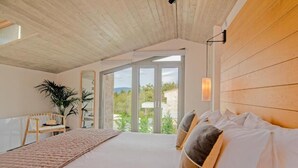 Ein Schlafzimmer mit einem großen Bett, Holzbalkendecke, Blick durch Glastüren, einem Sessel und einem Blumentopf.