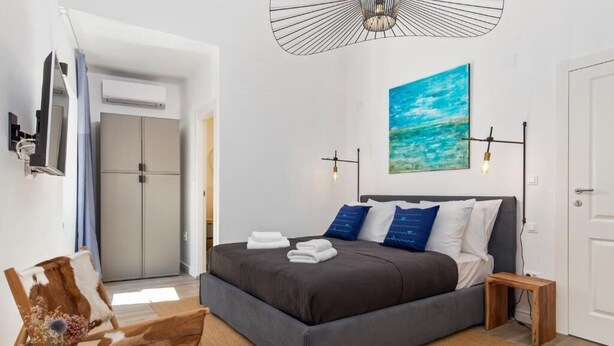 Un dormitorio moderno con una cama grande, una mesita de noche pequeña, un televisor montado en la pared y un cuadro de un cielo azul.