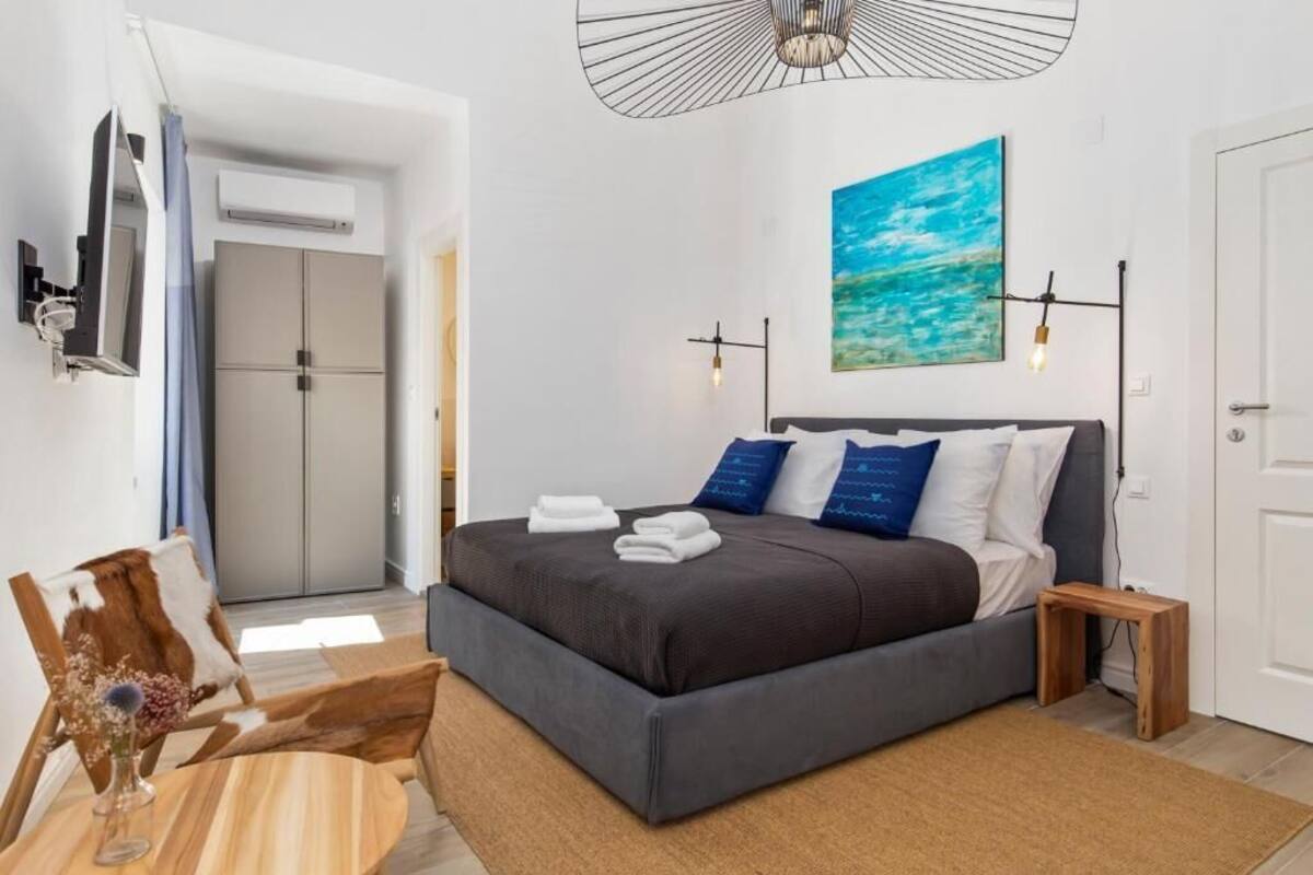 Un dormitorio moderno con una cama grande, una mesita de noche pequeña, un televisor montado en la pared y un cuadro de un cielo azul.