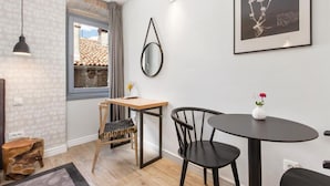 Una habitación moderna con una mesa de madera, dos sillas negras, una mesita redonda con un florero, un espejo colgado y una ventana con vistas a edificios.