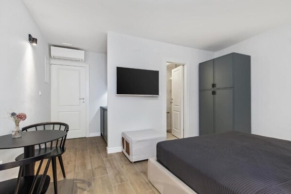 A compact room with a bed, a TV mounted on the wall, a small table with chairs, and a wardrobe.
