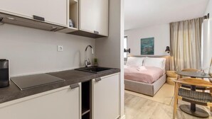 Una cocina moderna con estufa integrada, un lavamanos y un mueble pequeño. Hay una cama con una manta rosa y un cuadro en la pared.