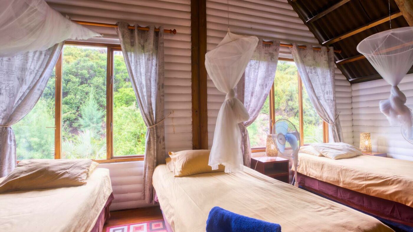 Una habitación con dos camas, un ventanal con cortinas, un ventilador de pie y una mesita de noche con lámpara.
