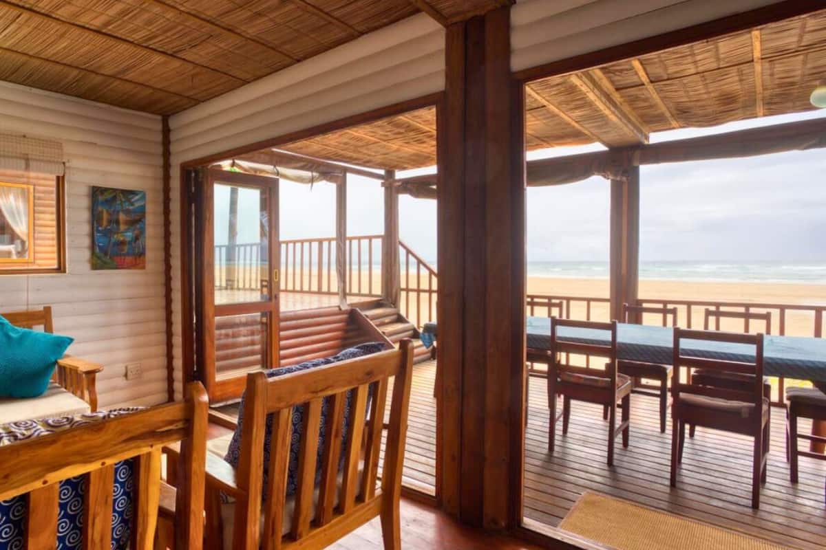A wooden deck with a wooden bench and table, a view of the beach, and a room with a patterned sofa and curtains.