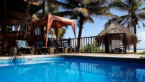 Un espace piscine avec des chaises longues, une structure à toit de chaume et des palmiers.