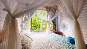 A bedroom with a canopy bed, a window with a view of greenery, and a bedside fan. - Farol da Barra (Inhambane)