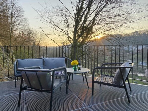 A balcony with a metal railing, a blue sofa, a round table with a vase of yellow flowers, and a view of trees and a sunset.