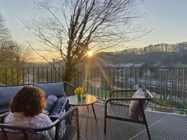 Un balcón con sofá y sillones, con vistas a un paisaje pintoresco con árboles y una lejana perspectiva.