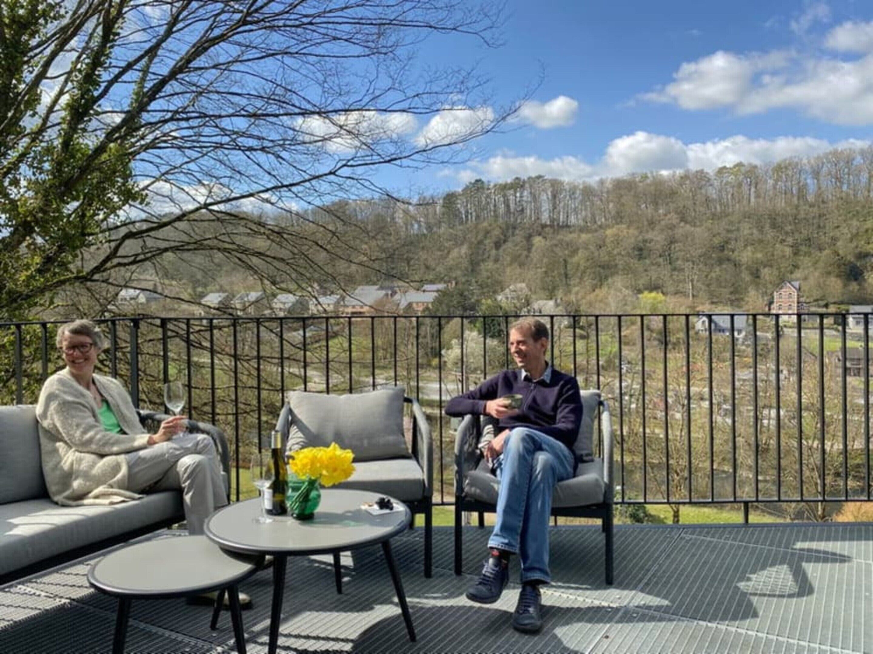 A terrace with two people seated on a grey sofa and chairs, a round table with a vase of yellow flowers, and a view of a forested landscape.