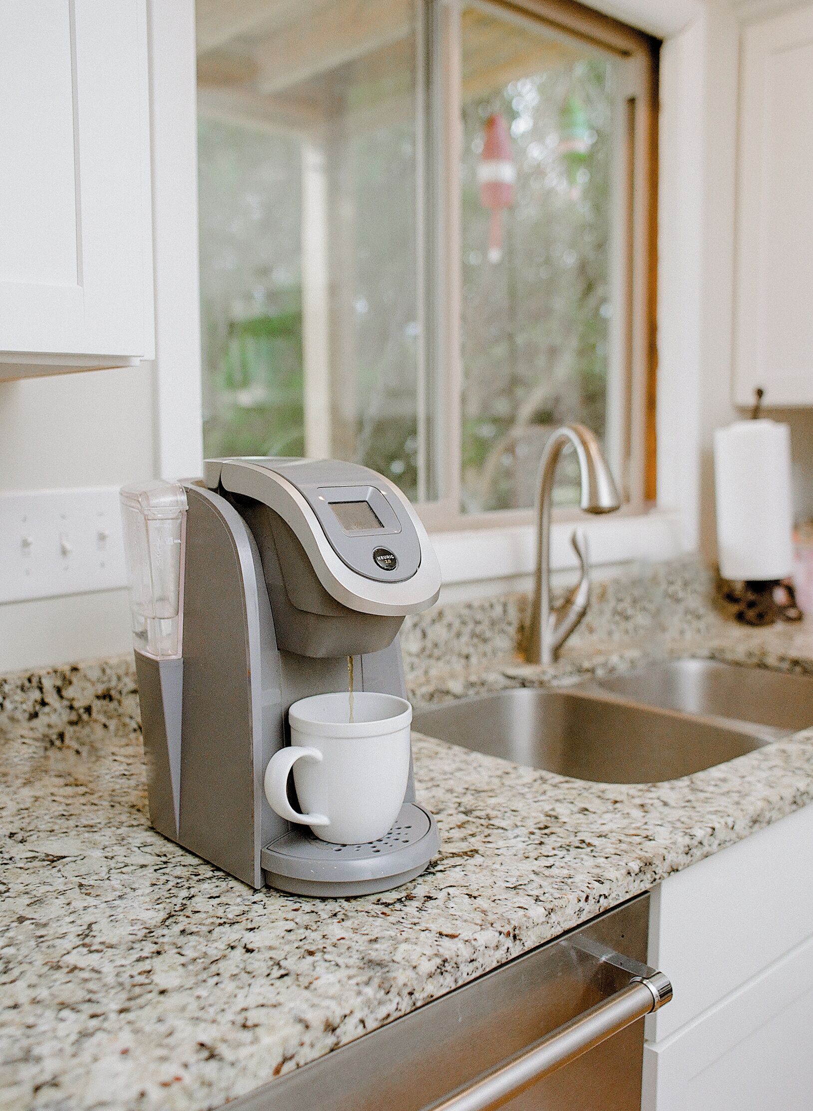 A coffee maker on a granite countertop with a window in the background.