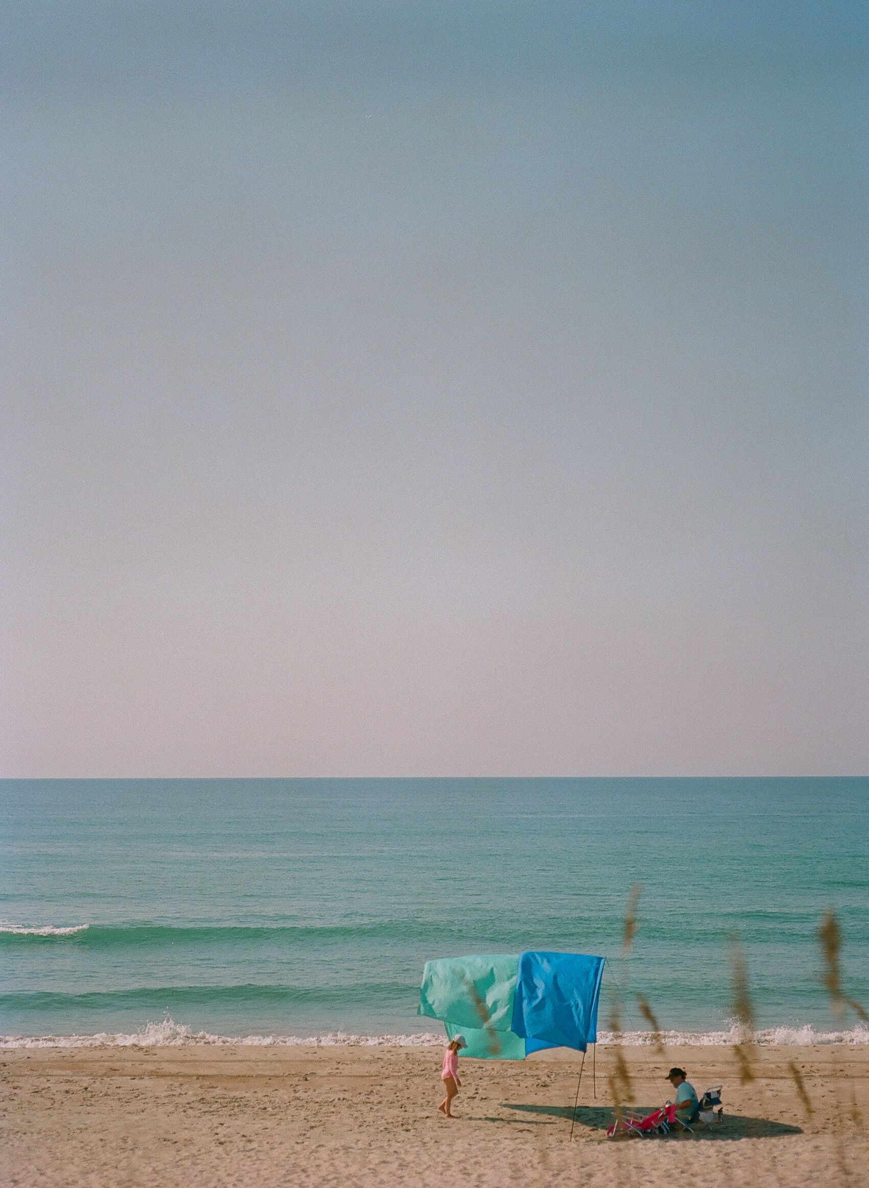 A beach with a blue umbrella and a person walking.