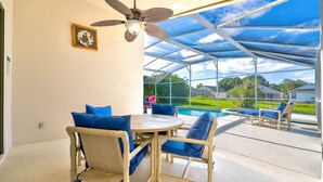 Un patio techado con una mesa y sillas, un ventilador de techo y vistas a una piscina y casas al fondo.