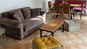 Ein Wohnzimmer mit einer Couch, einem Couchtisch, einem Rattan-Hocker, einem Esstisch mit Stühlen sowie einer Wand mit gerahmten Bildern und einem dekorativen Spiegel.