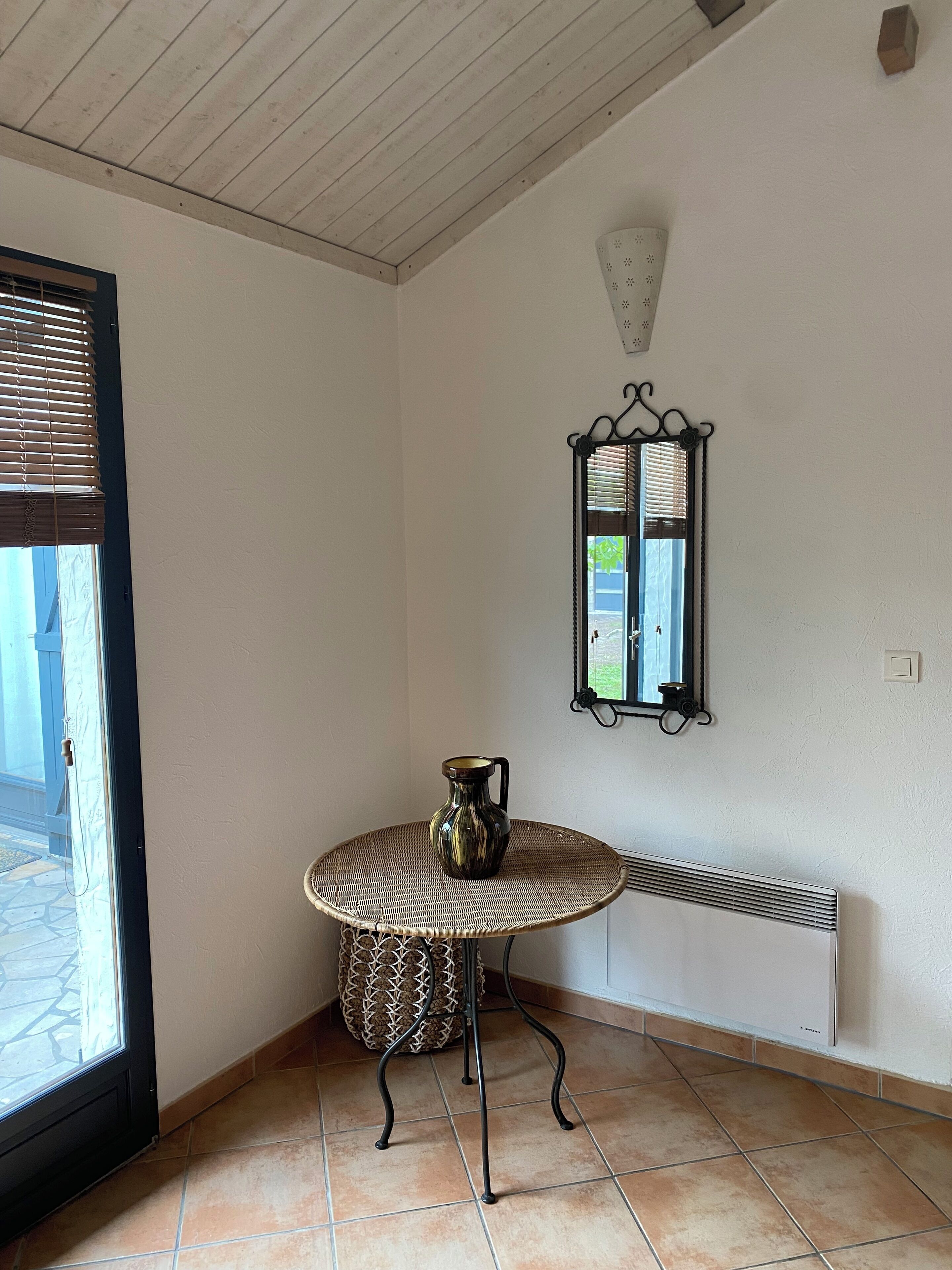 A corner room with a small round table, a decorative pitcher, a wall-mounted mirror, and a radiator.