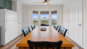 Une salle à manger avec une table et des chaises en bois, un ventilateur de plafond et de grandes fenêtres avec des rideaux.