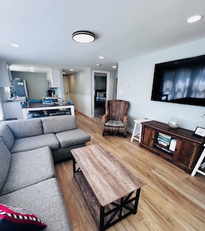 Uma sala de estar moderna com um sofá seccional, uma mesa de centro de madeira, uma poltrona de vime e uma TV de tela plana na parede.