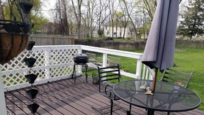A wooden deck with a round metal table and chairs, a barbecue grill, and hanging planters. - Musical Themed Duplex with Muskegon Lake Views (Muskegon)
