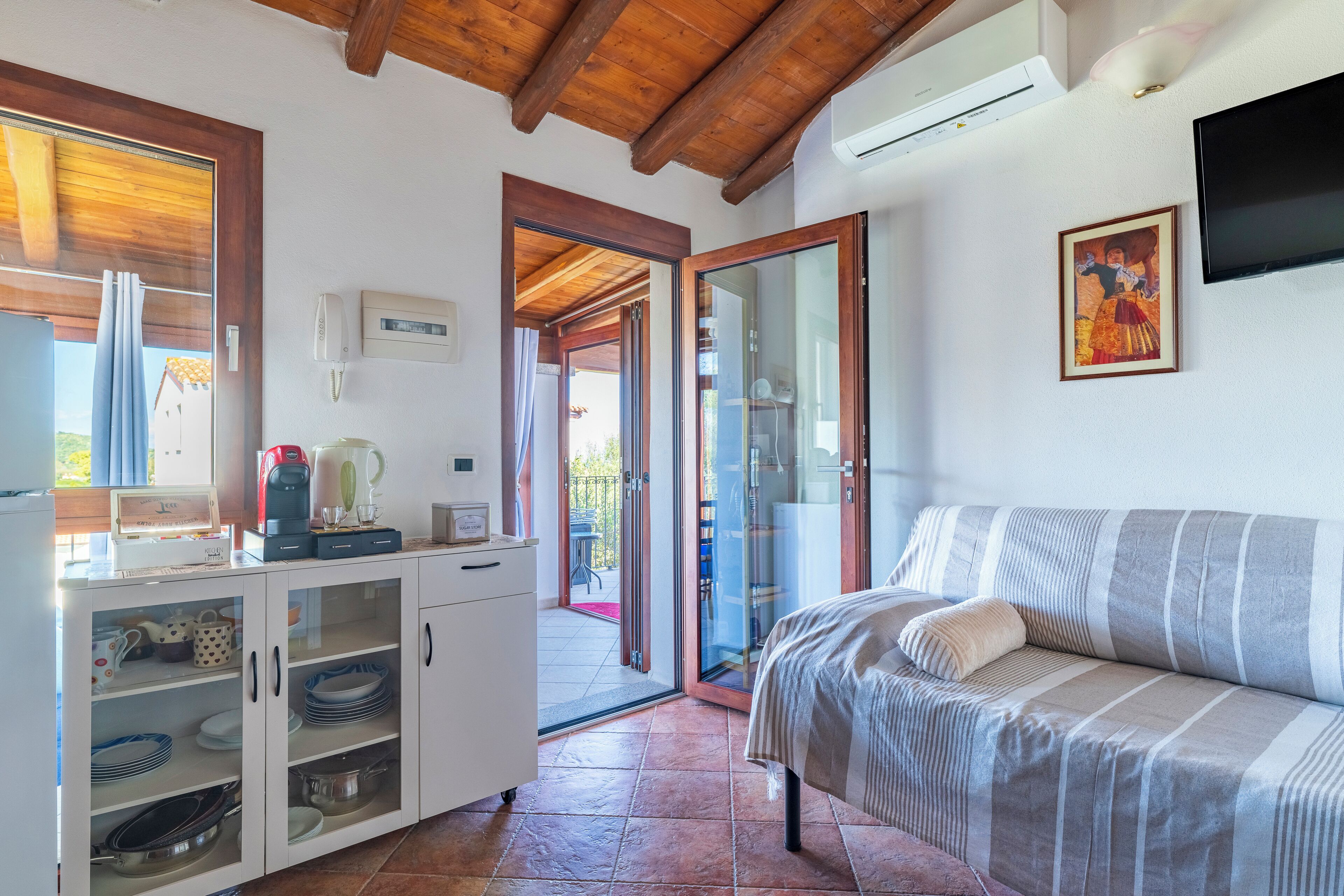 A compact room with a bed, a kitchenette with cabinets, a coffee maker, and a TV mounted on the wall.