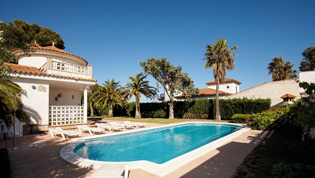 Ein Pool mit Liegestühlen, umgeben von Palmen und einem weißen Gebäude mit einem Ziegeldach.