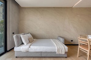 Une chambre à coucher moderne avec un grand lit, un petit canapé, une table à manger et une vue dégagée sur l’extérieur grâce à une grande fenêtre.