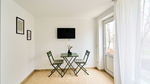 Una habitación pequeña con una mesa, dos sillas, un televisor de pantalla plana montado en la pared y una ventana con cortinas.