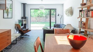 A modern living room with a dining table, chairs, a bookshelf, and a large window offering a view of the outdoors. - Mid-Century Modern Style 1BD House w/ Large Garden (Gloucestershire)
