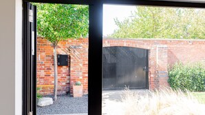 A view through a glass door into a courtyard with a brick wall, a black gate, and a small tree. - Mid-Century Modern Style 1BD House w/ Large Garden (Gloucestershire)