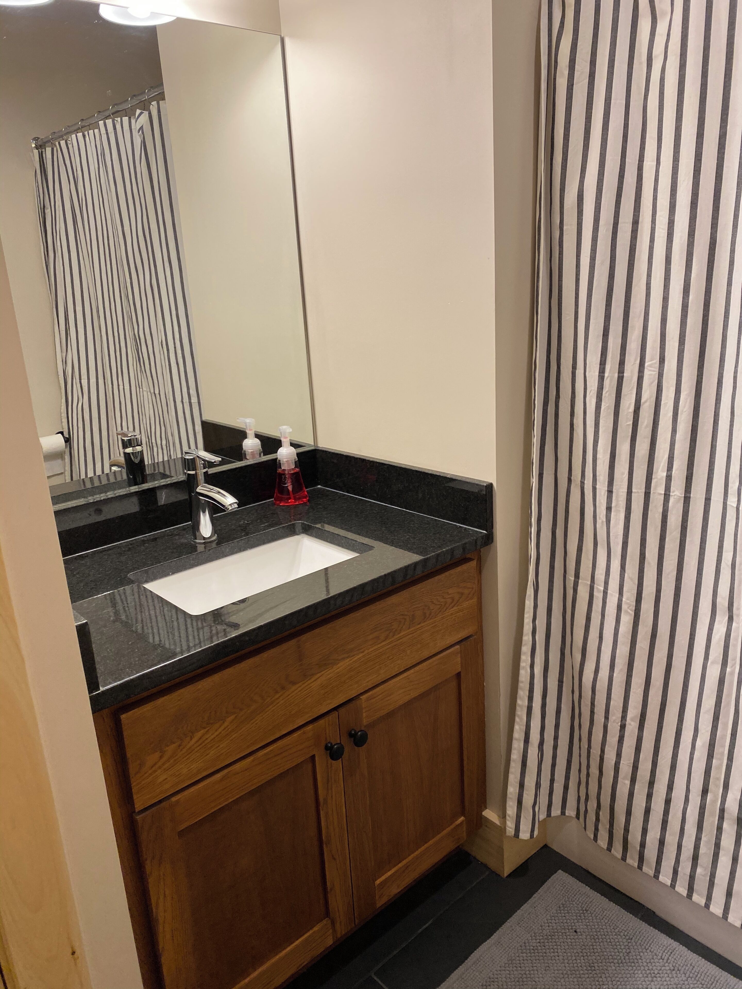 Un baño con encimera negra, lavabo blanco, armarios de madera y cortina de ducha a rayas.