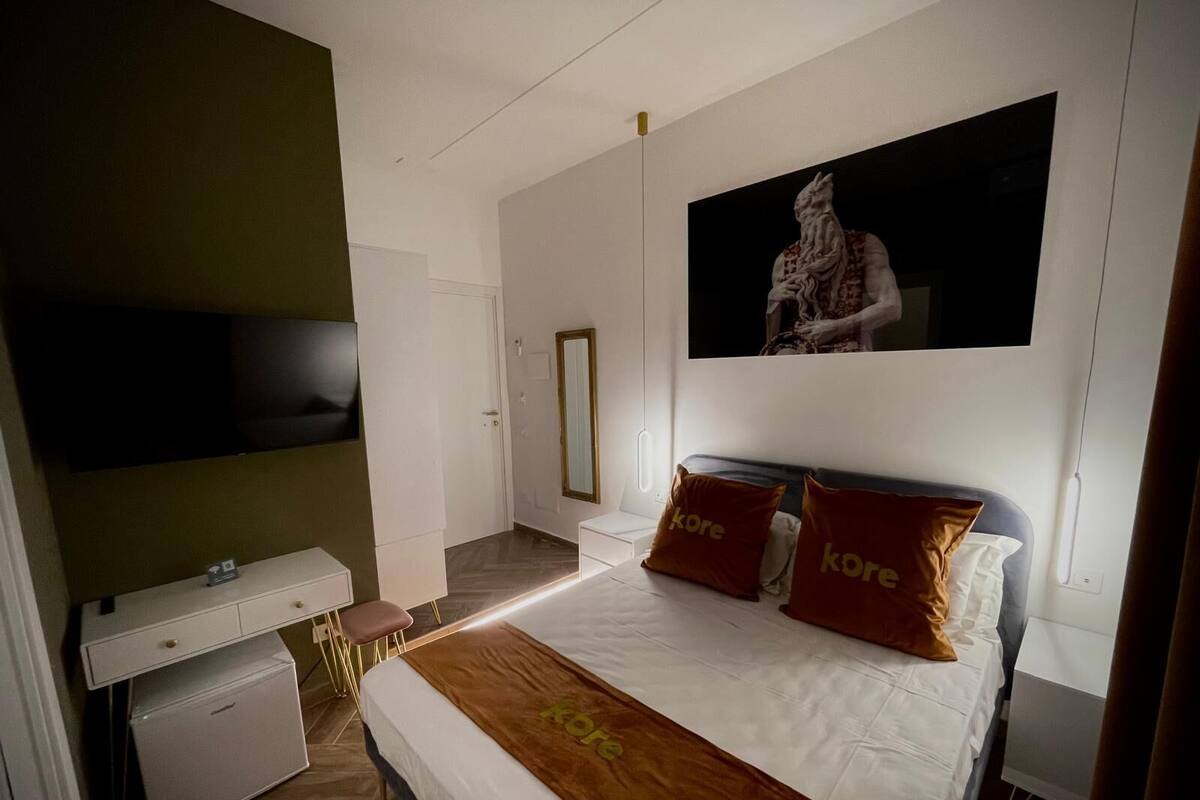 Una camera d'albergo moderna con un letto grande, una TV a schermo piatto montata a parete, una piccola scrivania con sedia e un frigorifero compatto.