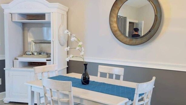 Un coin repas comprenant une table blanche, des chaises, un lustre, un miroir et un buffet.