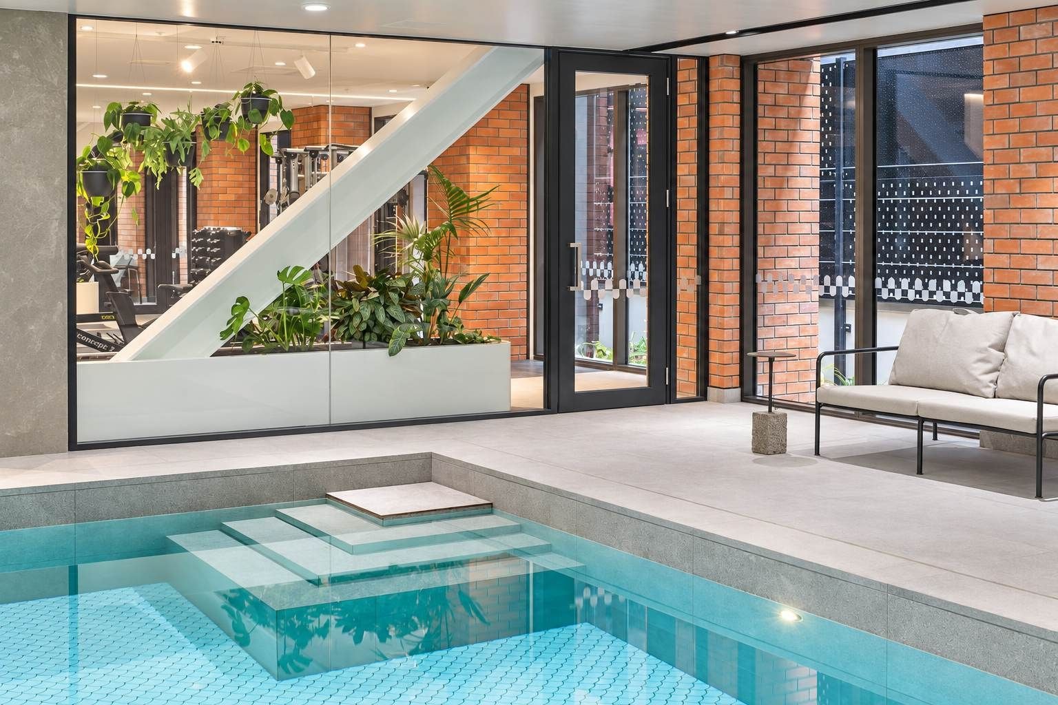 A modern indoor pool area with a glass barrier, a staircase, and a seating area with a sofa.