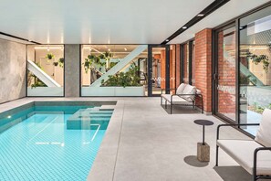 Un área de piscina interior moderna con una piscina azul cristalina, una escalera que conduce a un nivel superior y un espacio de descanso con un sofá y sillones.