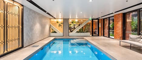 A modern indoor swimming pool with clear water, surrounded by a minimalist design with a staircase and seating area.