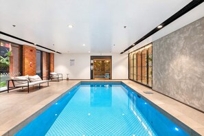 Una piscina interior moderna con superficie de agua azul cristalina, rodeada de sillones de descanso y un muro de vidrio.