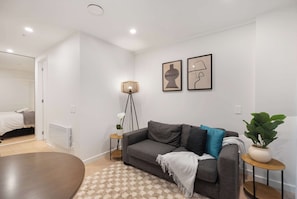 A modern living room with a grey sofa, a small side table with a lamp, a potted plant, and a patterned rug.