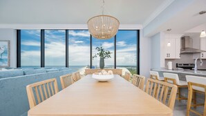 A modern dining area with a large wooden table, a bowl of fruit, and a view of the ocean through large windows.