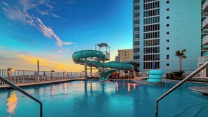 A swimming pool with a water slide, a tall building in the background, and a clear sky.