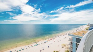A beach view from a high vantage point, featuring a clear sky, calm sea, and a sandy beach with umbrellas and people.