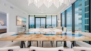 A modern living room with a large glass table, white furniture, and a view of the ocean.
