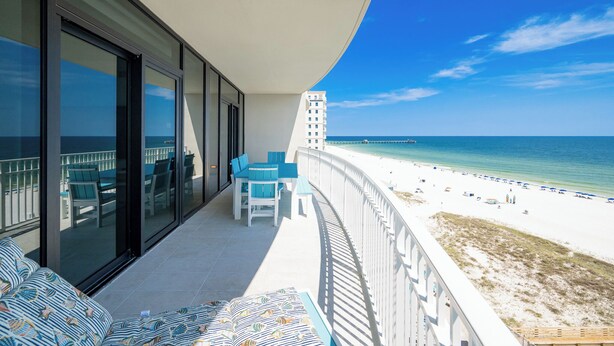 A balcony with a view of the beach, a pool, and a building in the background.