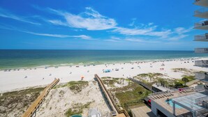A beach view from a balcony overlooking a sandy beach with a wooden boardwalk and a pool area.