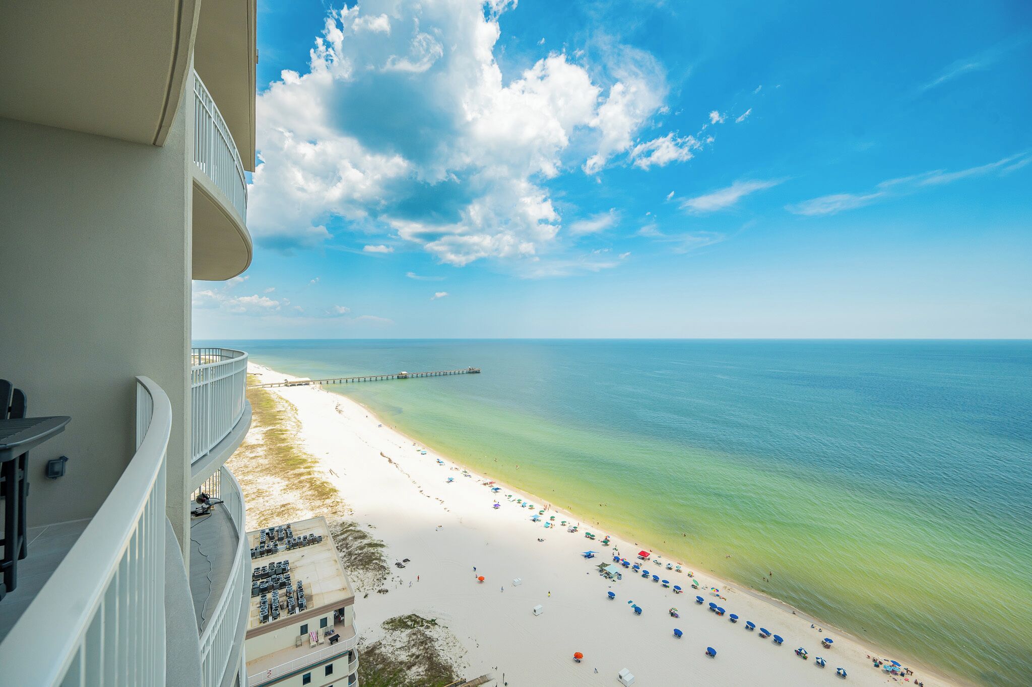 A beach view from a balcony with a clear sky, a sandy beach, and a pier in the distance.