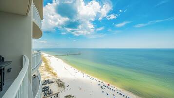A beach view from a balcony with a clear sky, a sandy beach, and a pier in the distance.