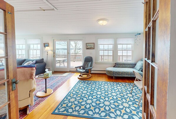 A cozy living room with a wooden floor, a patterned rug, a sofa, a chair, and a table with a lamp.