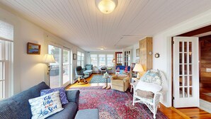 A spacious living room with a large window, a blue sofa, a white wicker chair, and a patterned rug. - Gorgeous Waterfront Home in the Heart of North Kingstown (North Kingstown)