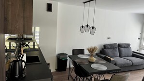 Una cocina moderna con estufa integrada, cafetera y cubo de basura. Hay un comedor con mesa, sillas y un sofá gris.