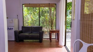 A room with a black leather sofa, a small wooden table, and a refrigerator. There is a large window with a view of greenery outside.